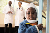 Young Kenyan boy looks closely at camera, two male medical workers in white coats stand in background