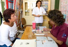 In clinic woman medical worker seated at table writes information on chart, woman across watches, another medical worker waits t