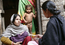 Woman holds swaddled baby in her arms, listening to woman across from her who has back to camera, another young child looks on