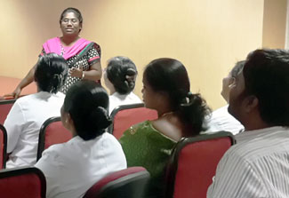 Faith Beulah speaks at the front of a classroom, many women and men sit in rows of chairs in the audience