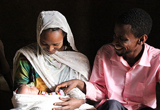 The photo captures an Ethiopian woman sitting beside her husband and holding her healthy newborn baby. The woman wears a white head scarf that also serves as a wrap, her husband a pink shirt. Both smile with delight.