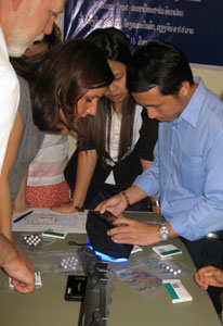 Group gathers around table, man uses black electronic handheld device to scan pills, more pills scattered on tabletop