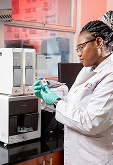 Female researcher working wearing lab coat and gloves examines testing supplies in front of lab equipment in a CAPRISA lab.