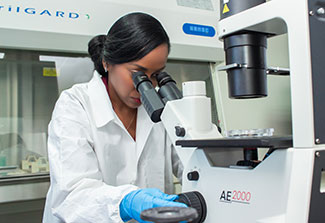 Dr. Simone Badal dressed in a lab coat looks in a microscope.