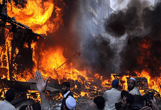 Bright orange flames surrounded by dark black smoke in a city street, buildings in background, people in foreground.