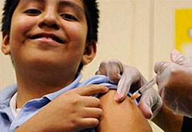 Boy receives shot in arm as he holds up his rolled sleeve, shot administered by clinician wearing gloves off camera