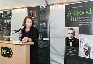 Mary Fogarty McAndrew speaks into a microphone in reception tent, displays with photos of Congressman John Fogarty in background