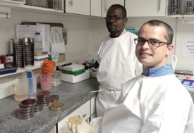 Brian Barnett seated in lab, holds lab dish, another man by his side, both looking at camera