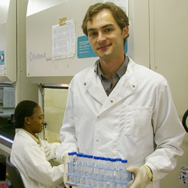 Ryan Davis in white lab coat wearing gloves holds many vials in holder, seated woman works at station in background