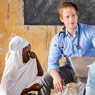 Dr. Cameron Gaskill, with a stethoscope around his neck, sits next to a young child wearing a headscarf