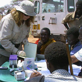 Woman researcher writes in notebook, people seated with blood sample collection supplies at outdoor table around her