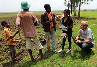 Two global health students collect data on mobile devices from community members, rural farmland in the background.