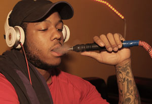 Young man exhales smoke after inhaling from hose of a waterpipe, wearing large headphones and baseball cap.