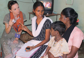 Lucy Horton holds container of medicine, looking at Indian woman holding child in lap, woman in between points to the bottle