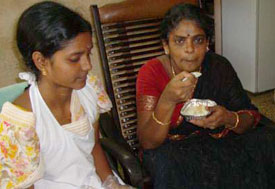 Indian woman, seated, moves spoonful of rice to her mouth, another woman in apron seated next to her observes