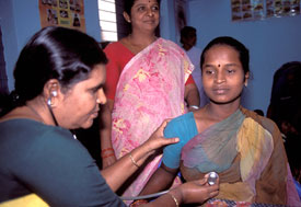 Indian woman seated in health clinic, female health care worker uses stethoscope to listen to chest