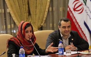 Woman wearing head scarf and man seated at conference table, woman speaks into microphone, Iranian flag in background