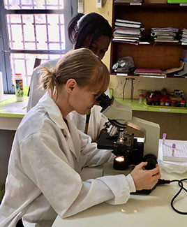 In a lab in Cameroon, Jillian Armstrong uses a microscope while another lab worker looks on.