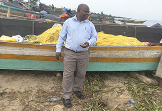 Dr. Joseph K.B. Matovu stands in front of a fishing boat on the shoreline in Uganda, many boats in the background.