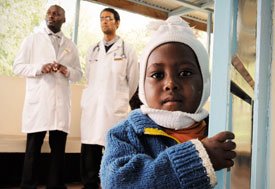 Young Kenyan boy looks closely at camera, two male medical workers in white coats stand in background