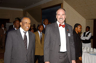 Botswana’s President Festus Mogae (left), wearing a pin striped suit and matching tie, meets Dr. Peter Kilmarx (right), wearing a grey suit and red bowtie at the 10th Anniversary of the CDC in Botswana in 2005. Behind them, you can see other attendees of this event.