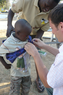 Outdoors, there is a man in a checkered shirt helps adjust a blue garment on a young child, who is being held by another person in the background.