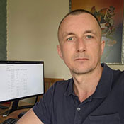 Dr. Kostyantyn Dumchev, wearing a black polo shirt, turns to take a selfie as he sits working on a computer in his office. A spreadsheet is visible on the computer screen behind him.