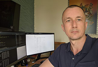 Dr. Kostyantyn Dumchev, wearing a black polo shirt, turns to take a selfie as he sits working on a computer in his office. A spreadsheet is visible on the computer screen behind him.