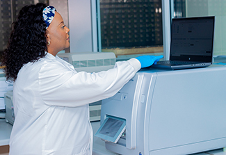 Photo of Catherine Koofhethile wearing a lab coat, working in a lab, typing on a laptop.