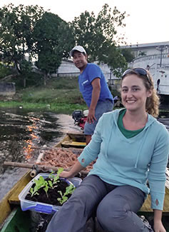 Dr. Leann Andrews seated in small boat on the water next to seedlings in soil, man steers from back of boat.