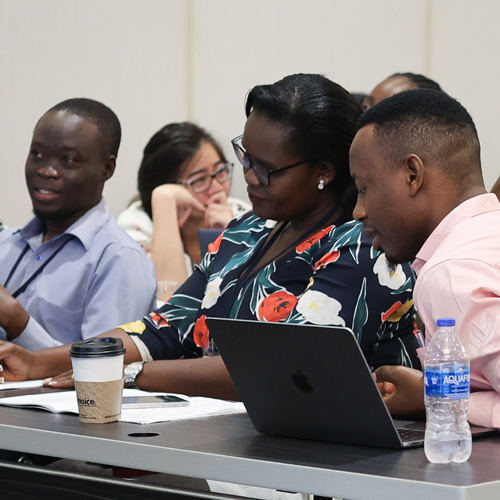A woman in a navy floral blouse and glasses leans in to look at a laptop screen with a man in a pink shirt seated beside her,