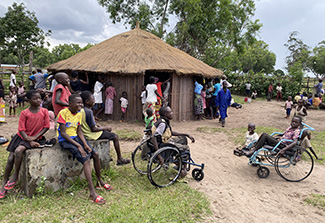 Adults and children gather near the thatched-roofed health center (in the background) in the village of Kahemba. In the foreground, a group of children – many of them smiling – sit on a cement block, on the ground, or in wheelchairs.
