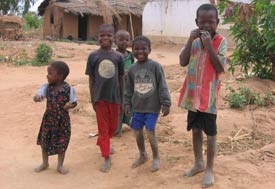 Four young boys and one girl, all barefoot on dusty ground, pose for camera in Malawi, hut in background