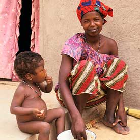 Woman in bright head wrap and clothing seated, smiling at camera on step next to naked toddler who is eating