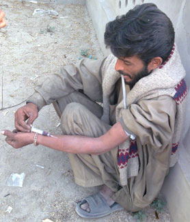 Pakistani man squats on ground against wall while injecting needle into forearm