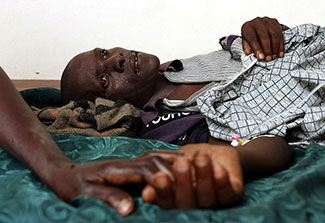 Man lying down in visible pain, his hand is held, in a hospital in Rakai, Uganda.