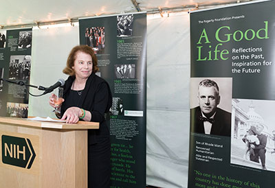 Mary Fogarty McAndrew speaks into a microphone in reception tent, displays with photos of Congressman John Fogarty in background