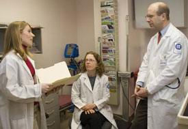 Three medical professionals in white lab coats, one holds open folder while speaking, other two listen