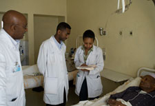 Three clinical workers in white jackets consulting notes stand next to patient in hospital bed