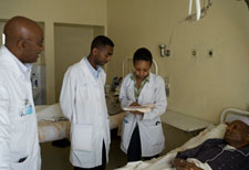 Three clinical workers in white jackets consulting notes stand next to patient in hospital bed