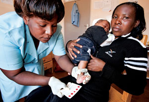 A young woman holds a baby on her lap, a medical worker examines the baby, referencing an informational card