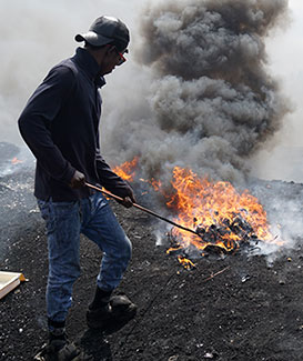 Man prods pile of burning electronic waste with a stick. Fire gives off a large cloud of thick, dark smoke.