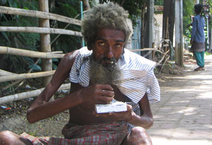 Emaciated man with scraggly beard and hair seated on the ground eats with his hands