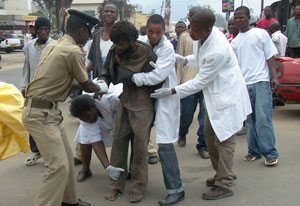 Three people in white lab coats and man in law enforcement uniform try to restrain a barefoot, disheveled man in a busy street