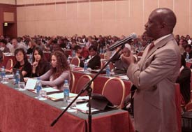 Conference room full of hundreds of people, one man in audience stands and speaks into microphone