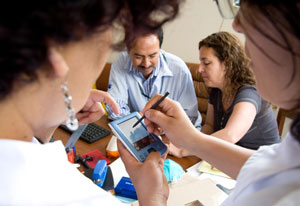 In foreground two medical workers view electronic device screen together, in background two more discuss together at a table