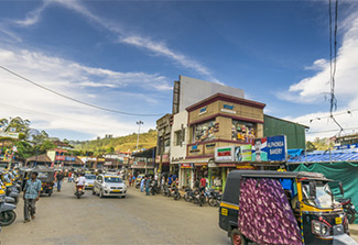 This photo shows a street in Kerala, India with cars, moto-taxis, bicycles, and people. A green hilltop is in the background.