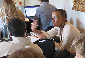 A woman wearing glasses and a light-colored blazer gestures with a pen while speaking, seated among a group of people in what appears to be a meeting or training session, with a computer monitor and framed picture visible in the background.