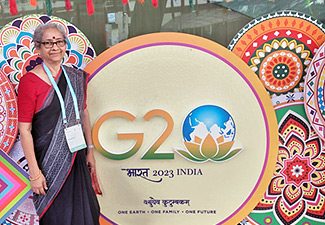 This photo shows Nandini Kumar wearing a red shirt and dark-colored sari, standing in front of a large rendering of the G20 2023 India logo.