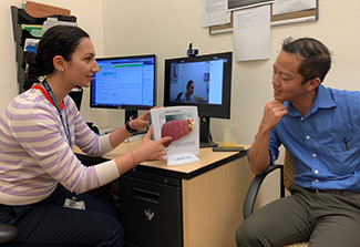 Dr. Nauzley Abedini seated at a desk in office, points to a model of a liver, discusses with person seated across from her.